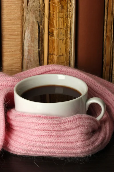 Cup of coffee wrapped in scarf on books background — Stock Photo, Image