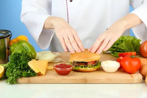 Mãos femininas preparando cheeseburger, sobre fundo azul — Fotografia de Stock