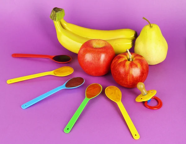 Purê de bebê em colheres com mamilo e frutas no fundo roxo — Fotografia de Stock