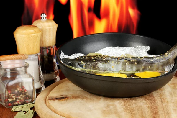 Fish in frying pan with herbs and lemon on board on wooden table on fire background — Stock Photo, Image