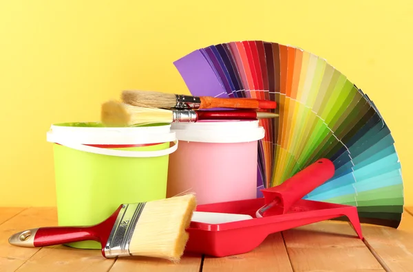 Paint pots, paintbrushes and coloured swatches on wooden table on yellow background — Stock Photo, Image