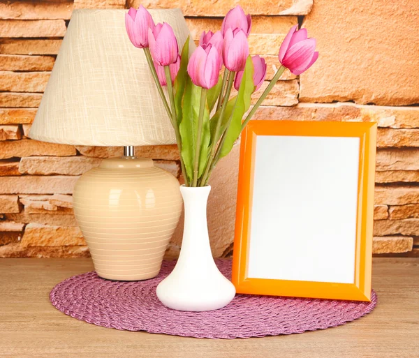 Красочная фоторамка, лампа и цветы на деревянном столе на фоне каменной стены — стоковое фото