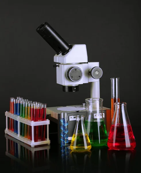Zkumavky s barevnými tekutinami a mikroskop na tmavě šedém pozadí — Stock fotografie