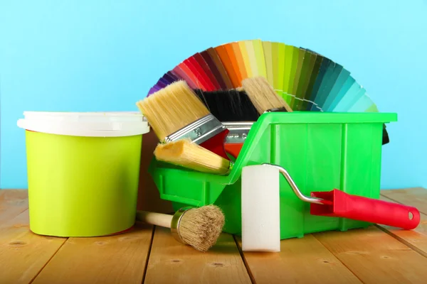 Paint pots, paintbrushes and coloured swatches on wooden table on blue background — Stock Photo, Image