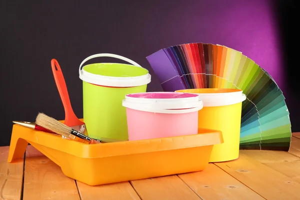 Paint pots, paintbrushes and coloured swatches on wooden table on dark purple background — Stock Photo, Image