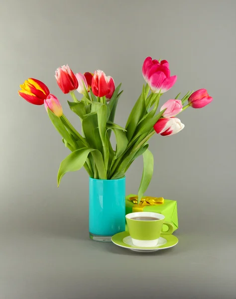 Piękne tulipany w wiaderku z darów i filiżankę herbaty na szarym tle — Zdjęcie stockowe