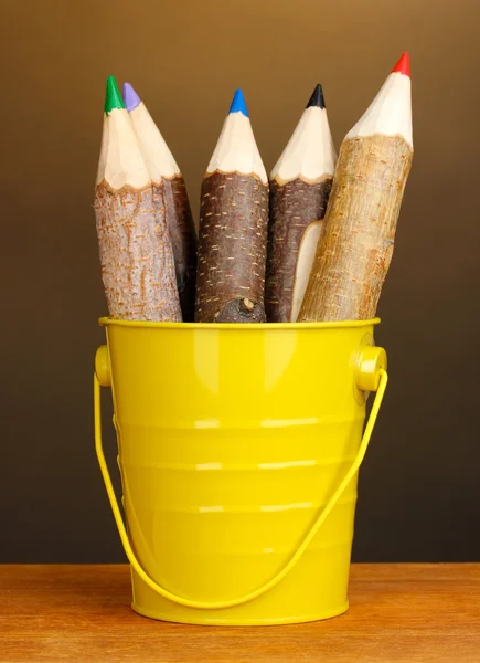 Цветные деревянные карандаши в желтом ведре на деревянном столе на коричневом фоне — стоковое фото