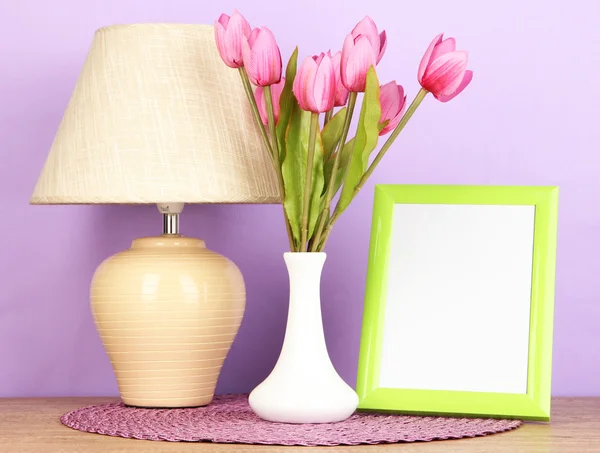 Moldura de foto colorida, lâmpada e flores na mesa de madeira no fundo lilás — Fotografia de Stock
