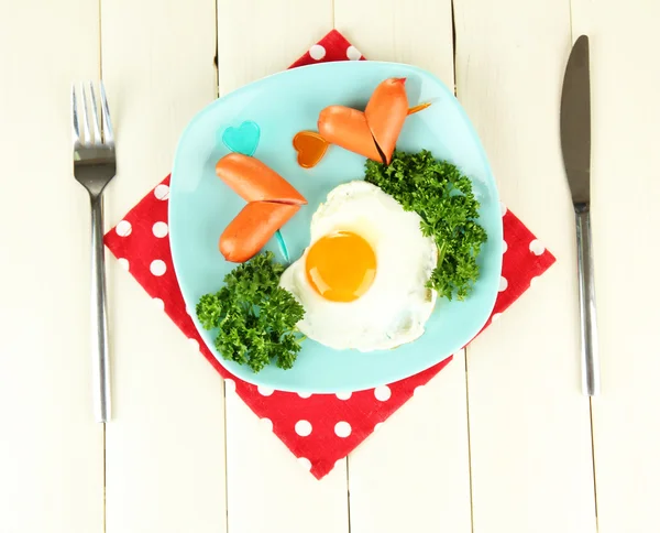 Сосиски у вигляді сердець, яєць і петрушки, на кольоровій тарілці, на дерев'яному фоні — стокове фото