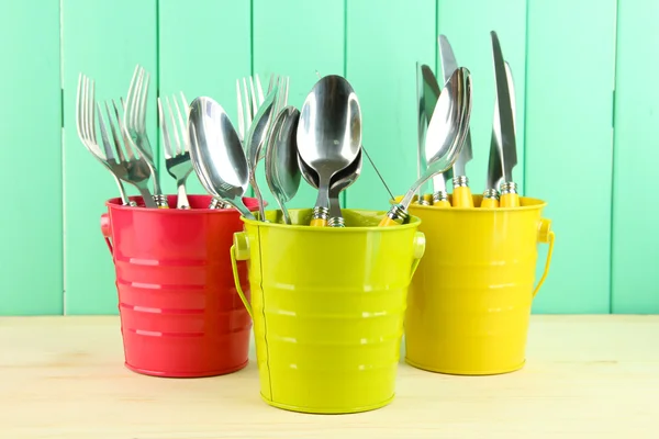 Knives, spoons, forks in colorful buckets on color wooden background — Stock Photo, Image