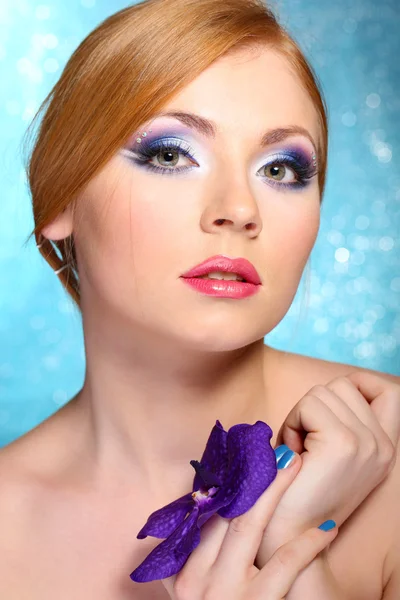 Hermosa mujer joven con glamour maquillaje y flor sobre fondo azul —  Fotos de Stock