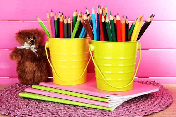 Colorful pencils in pails with copybooks and bear on table on pink background — Stock Photo, Image