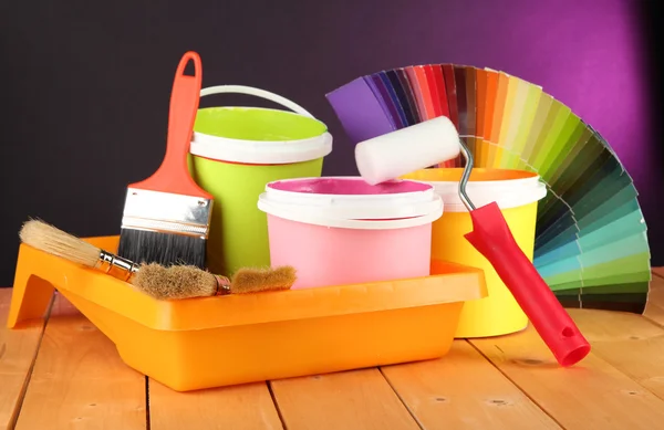 Paint pots, paintbrushes and coloured swatches on wooden table on dark purple background — Stock Photo, Image