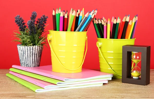 Kleurrijke potloden in twee emmers met copybooks op tafel op rode achtergrond — Stockfoto