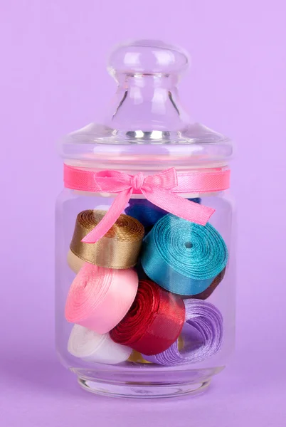 Glass jar containing various colored ribbons on lilac background — Stock Photo, Image