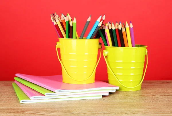 Crayons colorés dans deux seaux avec des copybooks sur la table sur fond rouge — Photo