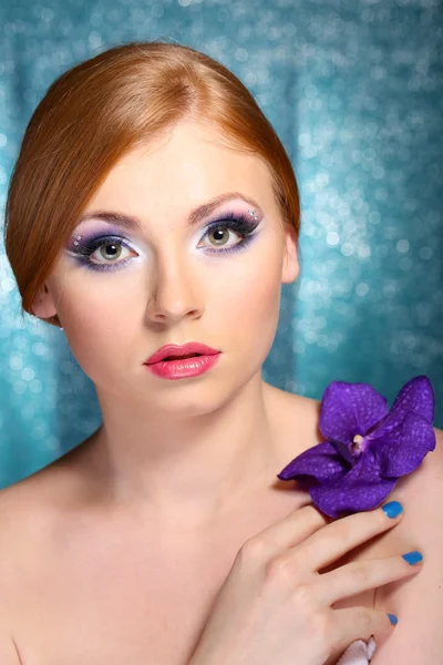 Hermosa mujer joven con glamour maquillaje y flor sobre fondo azul —  Fotos de Stock