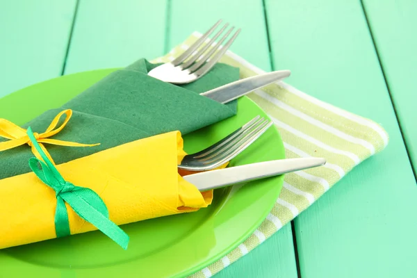 Вилки и ножи, завернутые в зеленые и желтые бумажные салфетки, на цветном деревянном фоне — стоковое фото