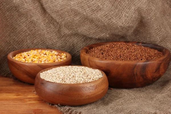 Raw corn,buckwheat and wheat in wooden bowls on table on sackcloth background — Stock Photo, Image