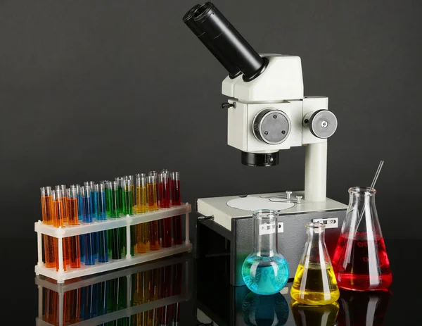 Δοκιμαστικοί σωλήνες με πολύχρωμο υγρών και μικροσκόπιο σε σκούρο γκρι φόντο — Φωτογραφία Αρχείου