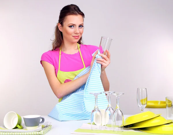 Mulher bonita limpa utensílios limpos na cozinha em fundo cinza — Fotografia de Stock