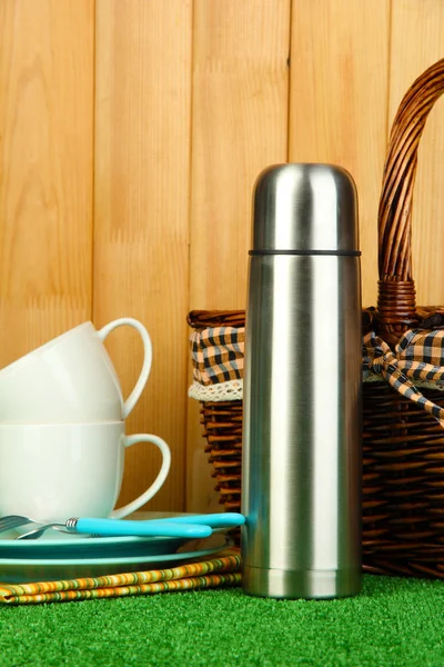 Metal thermos with cups, plates and basket on grass on wooden background — Stock Photo, Image