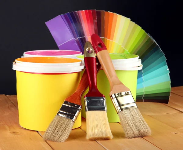 Paint pots, paintbrushes and coloured swatches on wooden table on dark yellow background — Stock Photo, Image