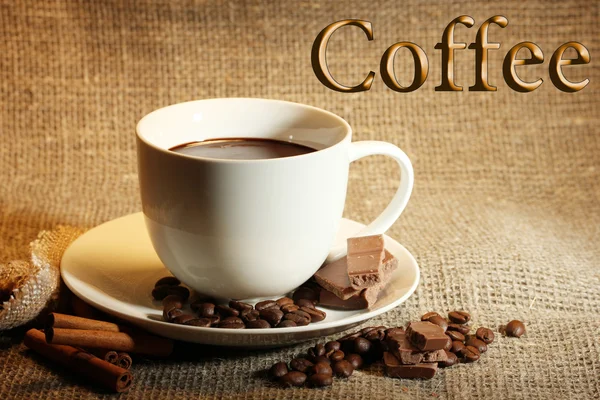 Kopp kaffe och bönor, kanelstänger och choklad på säckväv bakgrund — Stockfoto