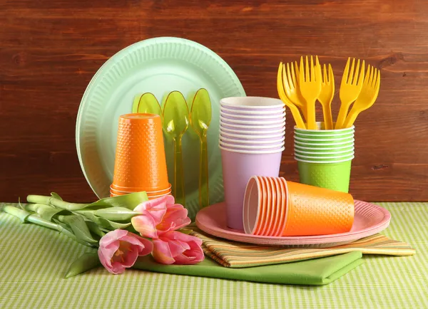 Multicolored plastic tableware on table with tulips on wooden background — Stock Photo, Image