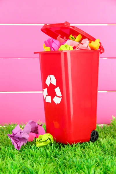 Papelera de reciclaje con papeles sobre hierba sobre fondo rosa — Foto de Stock
