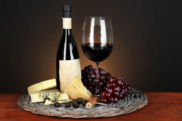 Refined still life of wine, cheese and grapes on wicker tray on wooden table on brown background — Stock Photo, Image