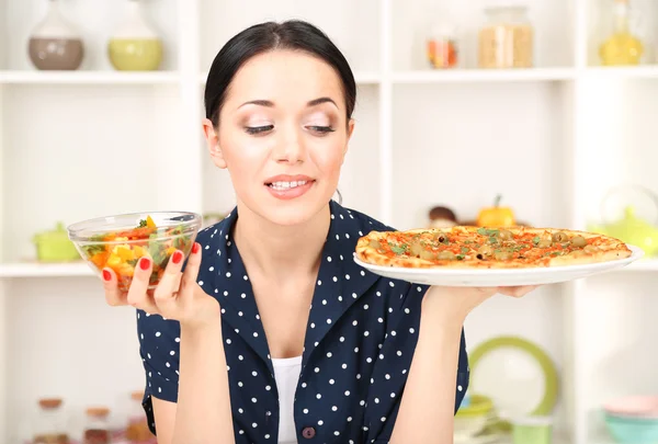 Pretty girl selects pizza or diet on kitchen background — Stock Photo, Image