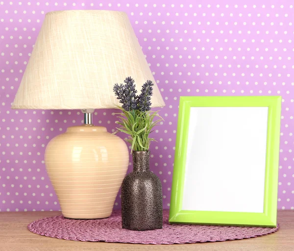 Colorful photo frame, lamp and flowers on wooden table on lilac polka dots background — Stock Photo, Image