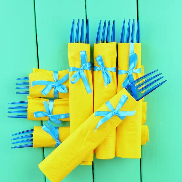 Голубые пластиковые вилки, завернутые в салфетки из желтой бумаги, на фоне цвета дерева — стоковое фото