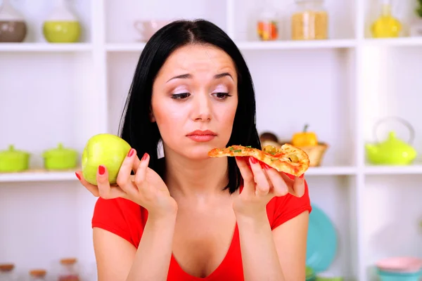 Pretty girl selects pizza or diet on kitchen background — Stock Photo, Image