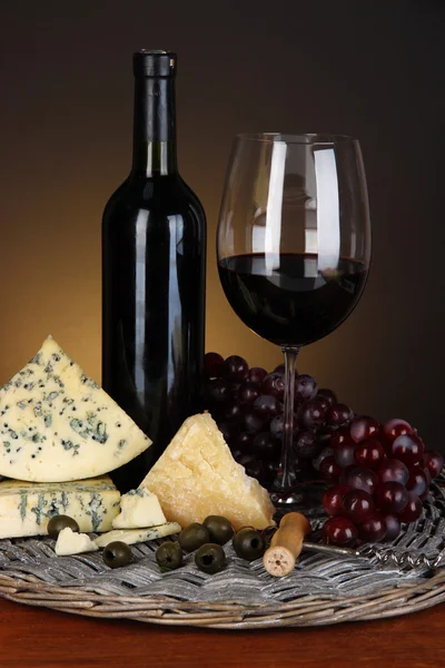 Refined still life of wine, cheese and grapes on wicker tray on wooden table on brown background — Stock Photo, Image