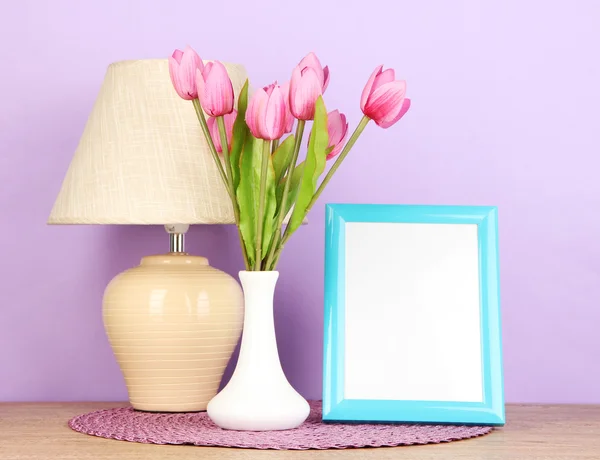 Colorful photo frame, lamp and flowers on wooden table on lilac background — Stock Photo, Image