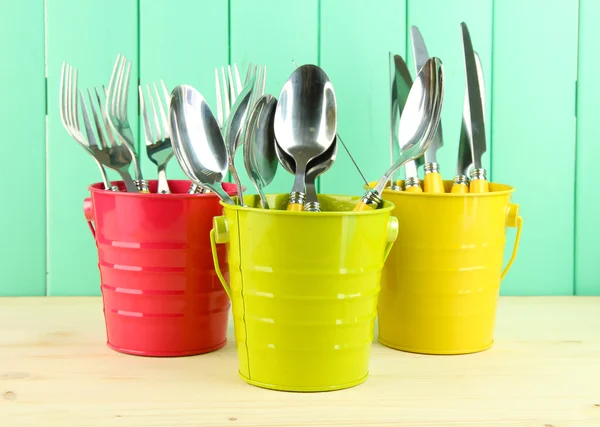 Knives, spoons, forks in colorful buckets on color wooden background — Stock Photo, Image