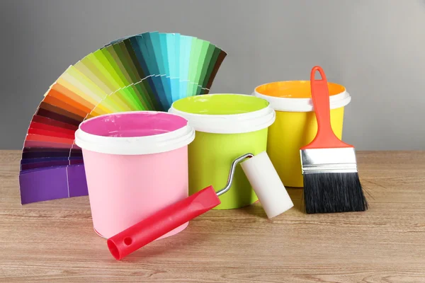 Paint pots, paintbrushes and coloured swatches on wooden table on grey background — Stock Photo, Image
