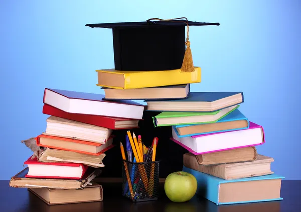 Livros e magister cap contra placa da escola em mesa de madeira no fundo azul — Fotografia de Stock