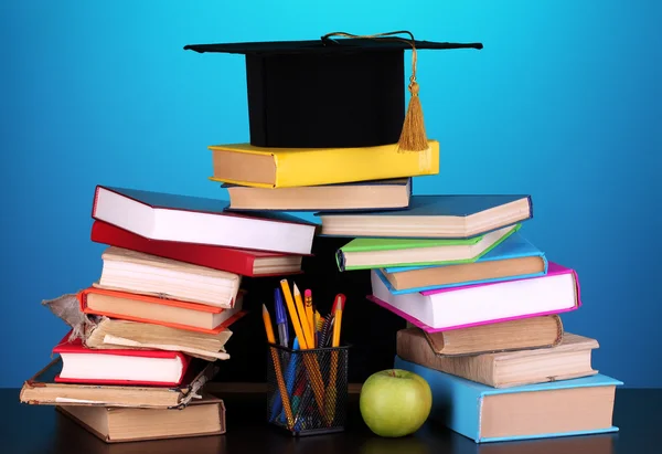 Books and magister cap against school board on wooden table on blue background — Stock Photo, Image