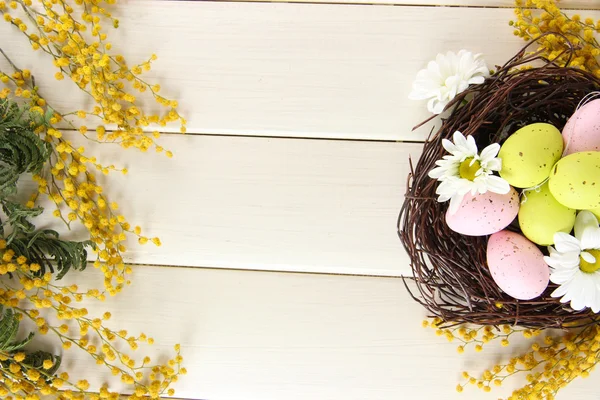 Uova di Pasqua in nido e fiori di mimosa, su fondo di legno bianco Foto Stock Royalty Free