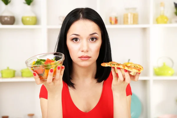 Mooi meisje selecteert pizza of dieet op keuken achtergrond — Stockfoto