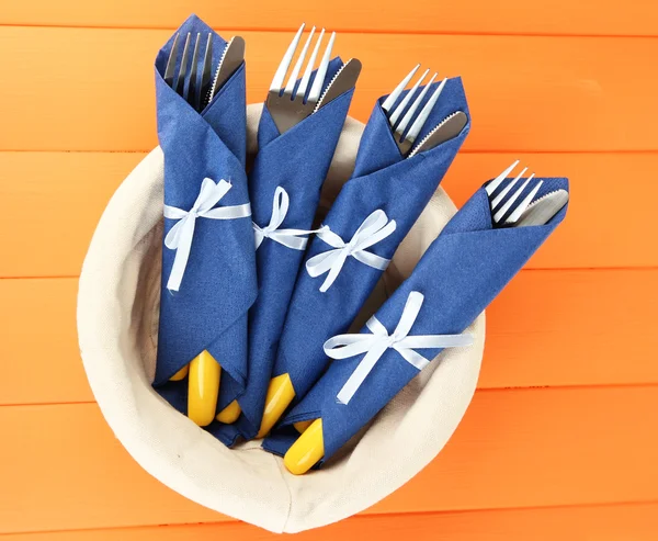 Tenedores y cuchillos envueltos en servilletas de papel azul, en cesta, sobre fondo de madera de color — Foto de Stock