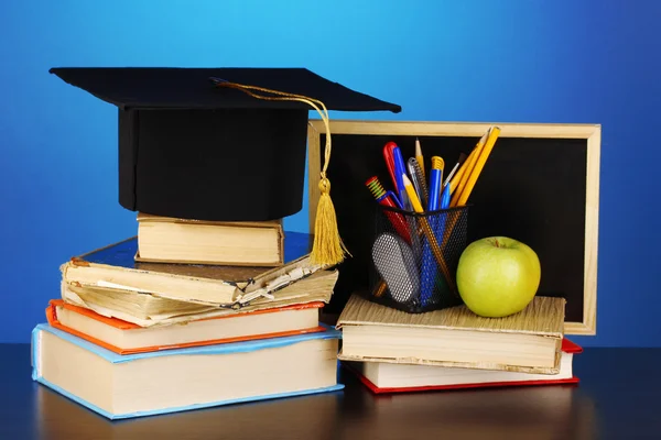 Books and magister cap against school board on wooden table on blue background — Stock Photo, Image