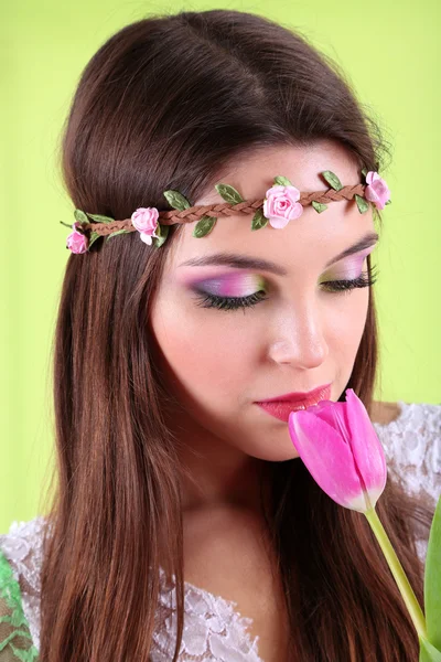 Young beautiful girl in decorative wreath with tulip in her hand, on green background — Stock Photo, Image