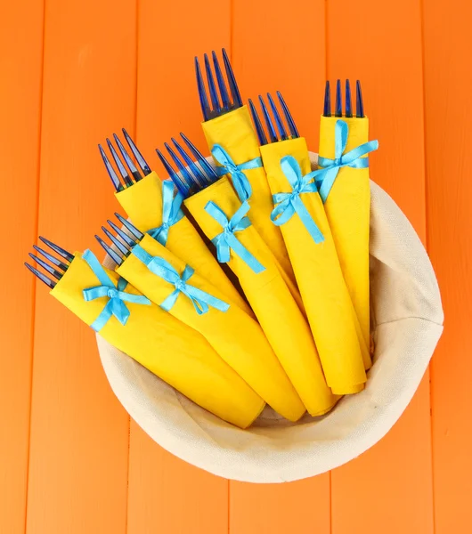 Keltaiseen paperipyyhkeeseen käärityt siniset muovihaarukat, korissa, värillisellä puupohjalla — kuvapankkivalokuva