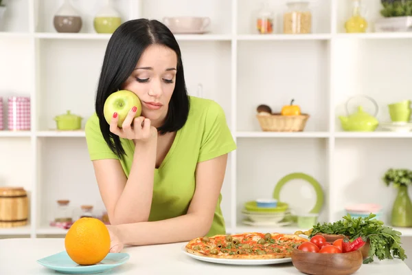 Mooi meisje selecteert pizza of dieet op keuken achtergrond — Stockfoto