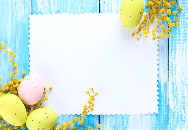 Пустая открытка с пасхальными яйцами и цветами мимозы на синем деревянном фоне — стоковое фото