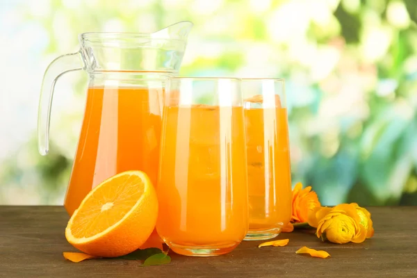 Glasses and pitcher of orange juice on wooden table, on green background — Stock Photo, Image